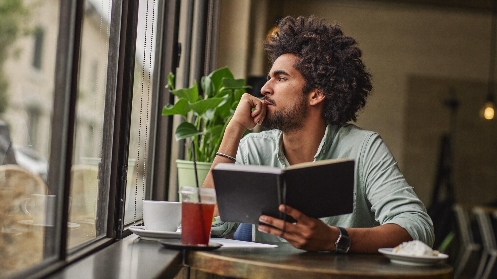 Man with full curly hair and a beard looking out of a cafe window with a notebook