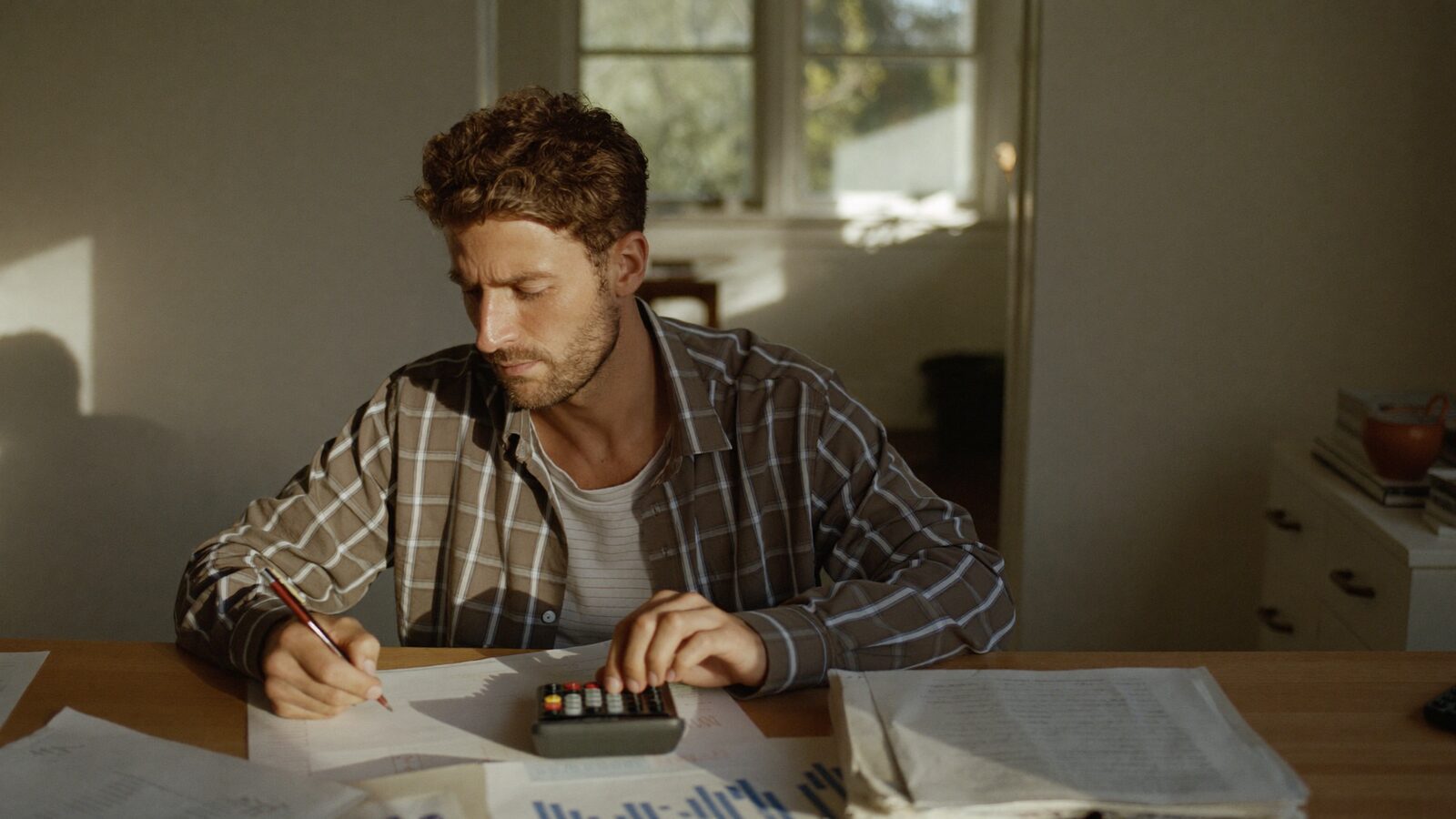 Man with full curly hair working through paperwork with a calculator at a home desk
