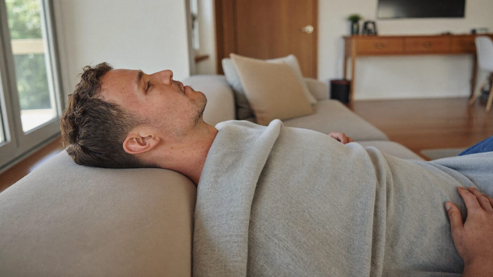 Man with short hair resting under a blanket on a sofa in a bright living room