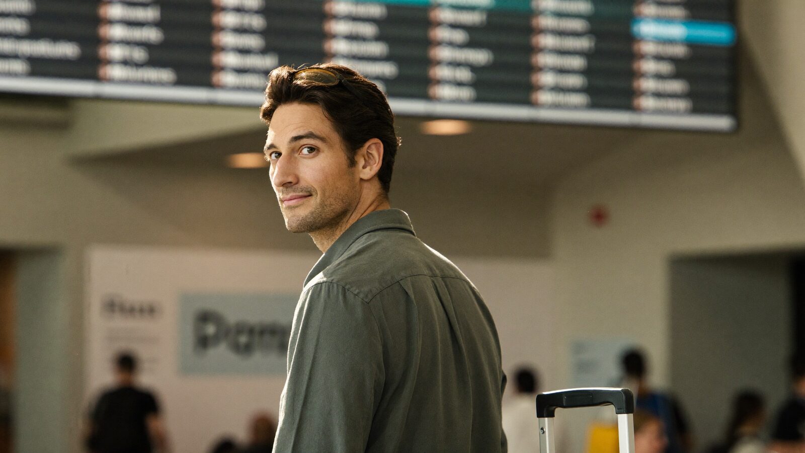 Man with full hair and a suitcase in an airport terminal in front of a departures board