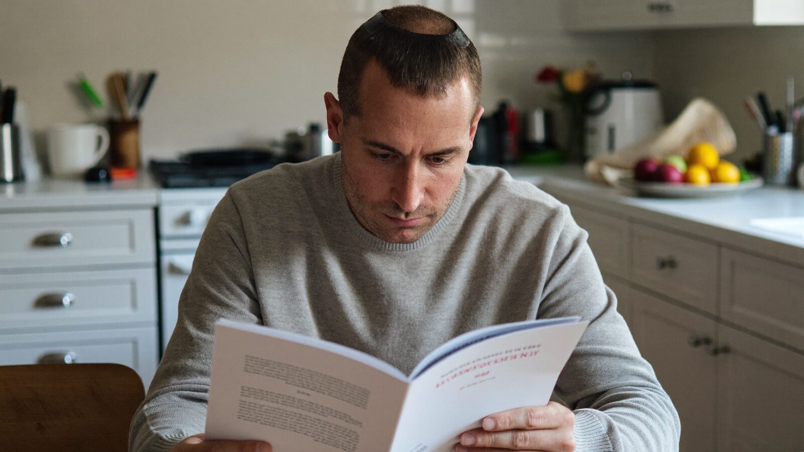 Man with short hair reading a printed brochure at a kitchen table