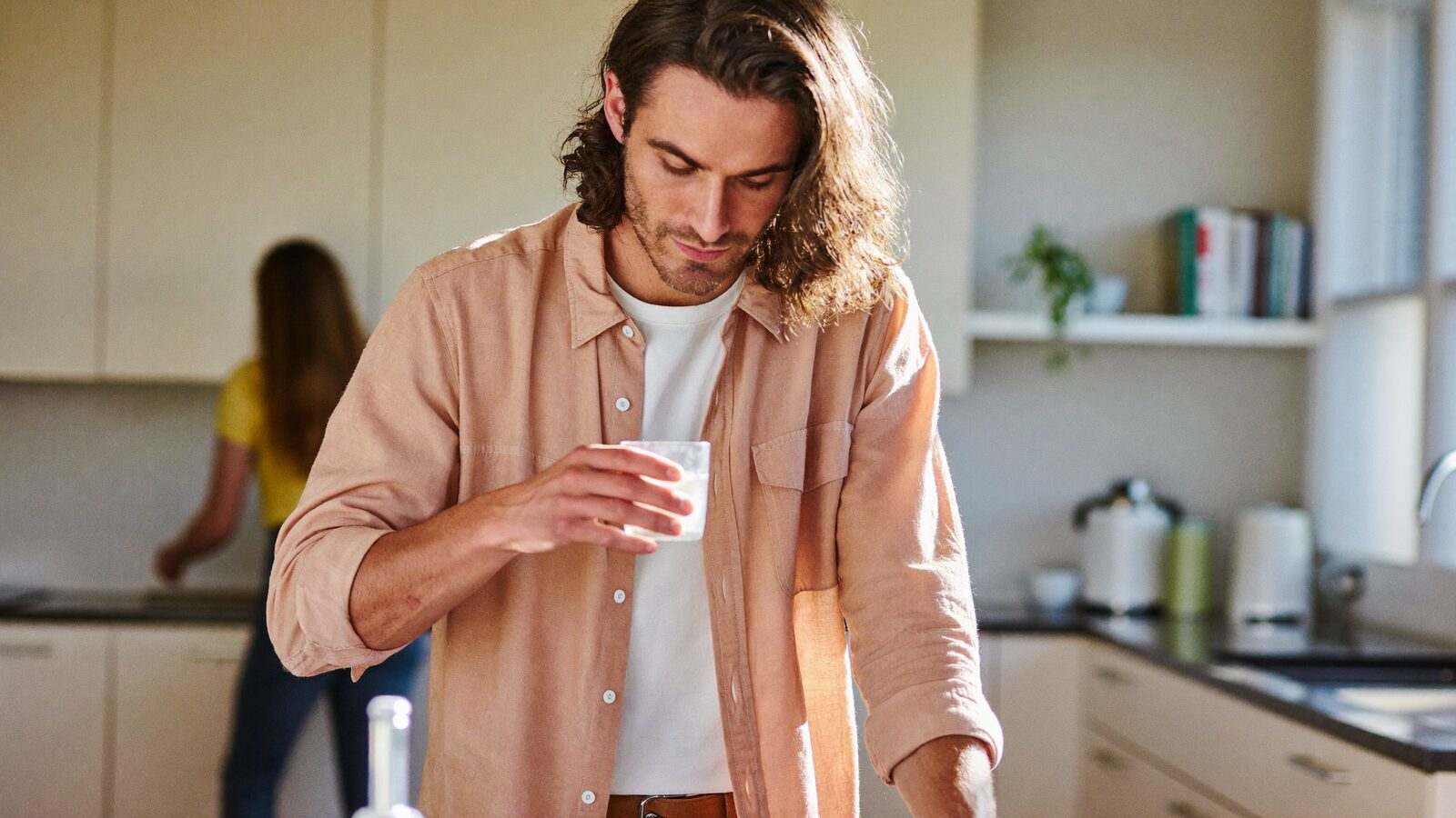 Man with long, thick wavy hair drinking a glass of water in a bright kitchen