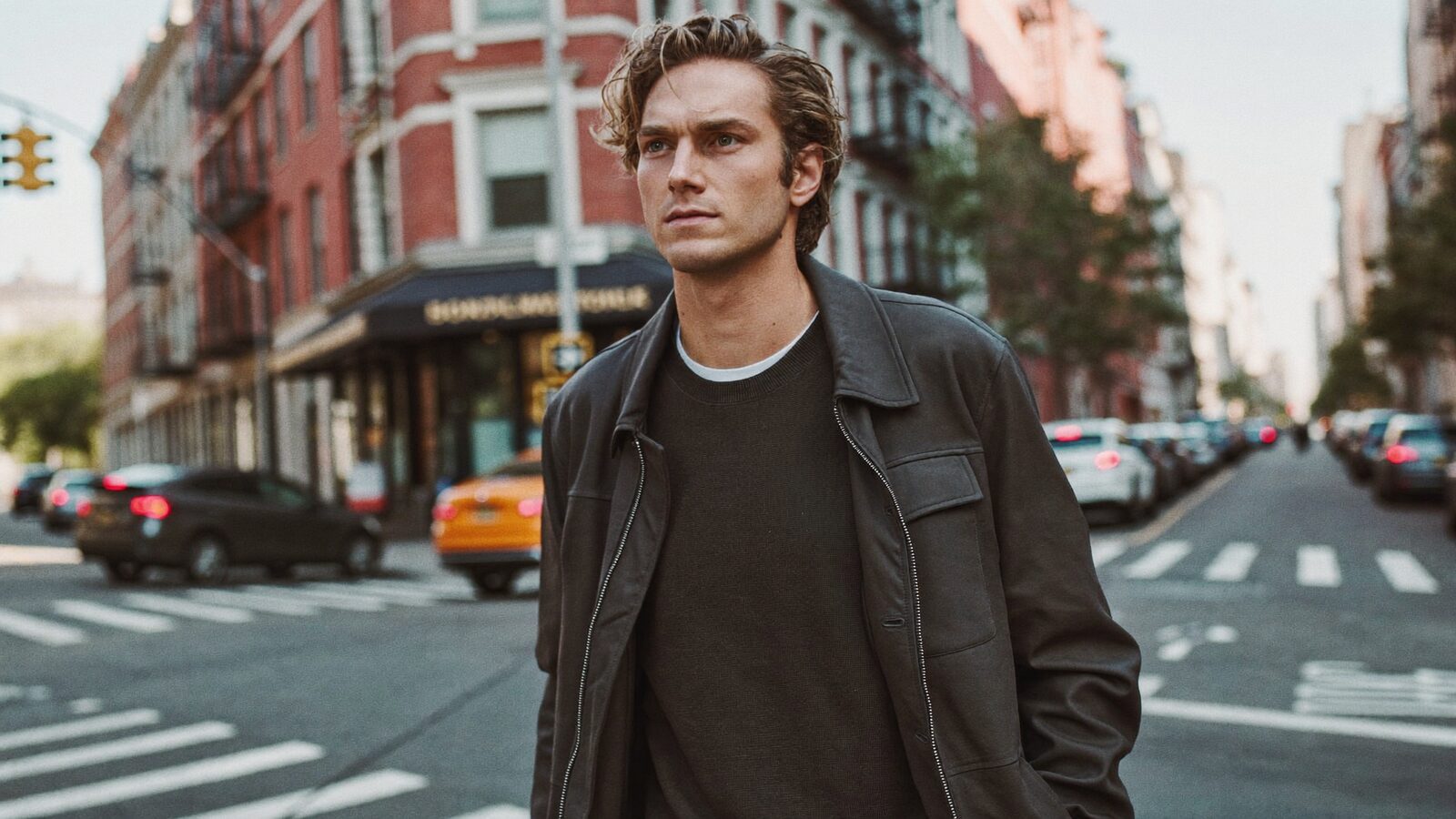 Man with full wavy hair in a dark jacket walking across a city street