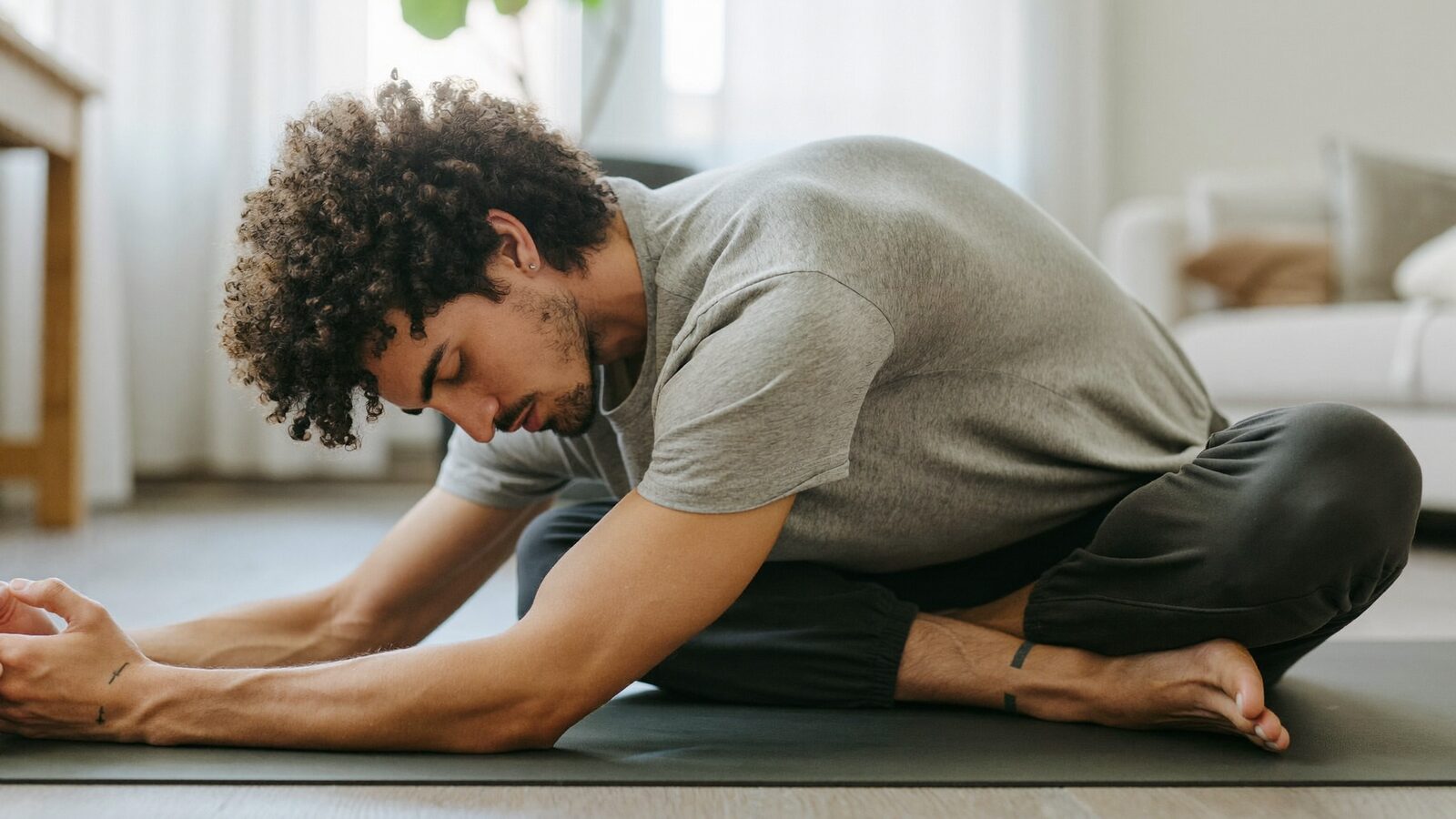 Man with thick curly hair in a gray t-shirt stretching on a yoga mat at home